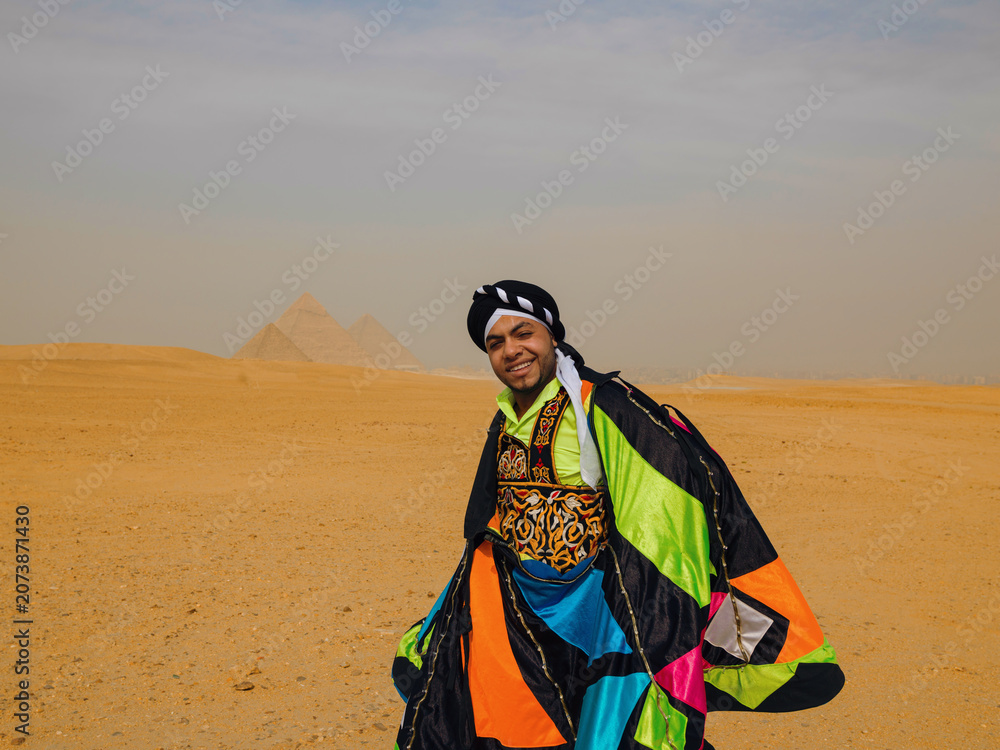 Fototapeta premium Joyful Egyptian tanoura dancer in vibrant multicolor folk costume smiling proudly in the desert before the Giza pyramids ideal for cultural festival