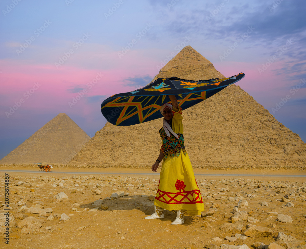 Fototapeta premium Vibrant Egyptian tanoura dancer twirling a colorful folklore skirt overhead before the Giza pyramids under a pink twilight sky ideal for Egypt travel