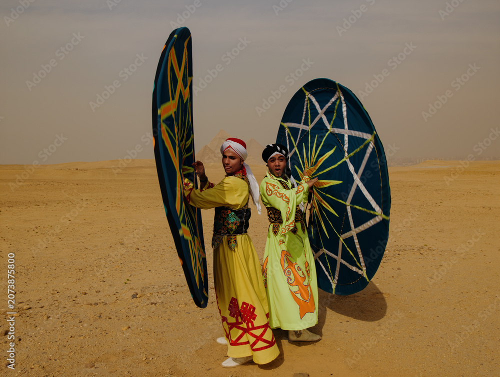 Fototapeta premium Two energetic Egyptian Tanoura folk dancers performing with colorful star patterned spinning skirts in the desert near the Giza pyramids ideal for