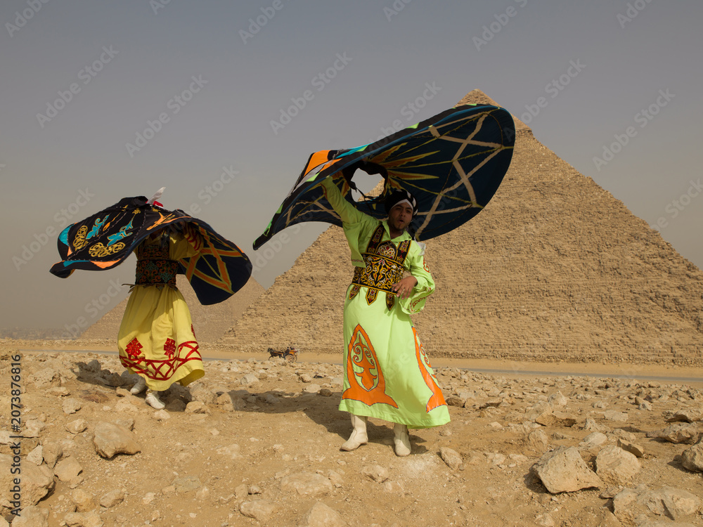 Fototapeta premium Two Egyptian Tanoura folk dancers spinning vibrant colorful skirts overhead in an energetic whirling performance before the ancient Giza pyramids