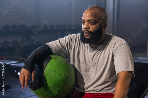 Fotografie African American man sitting on bench leaning on green medicine ball in gym, wea