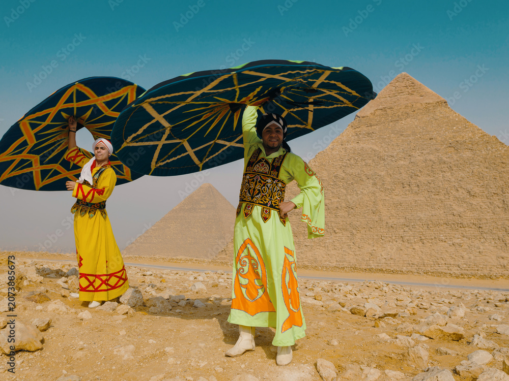 Fototapeta premium Two joyful Egyptian tanoura folk dancers spinning colorful decorated skirts overhead in bright desert sunlight before the Giza pyramids ideal for
