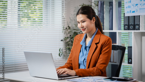 Smiling young businesswoman working on a laptop at her office desk, with reports, shelves and window blinds in the background, showing a calm and productive workday.