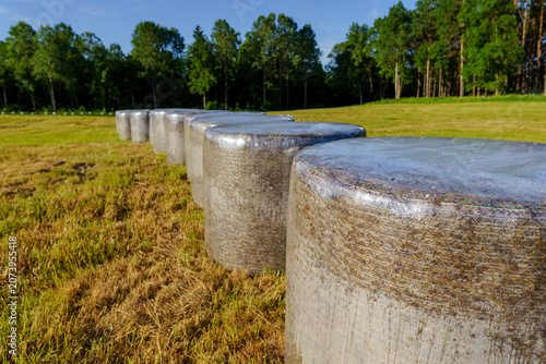 Scenic agricultural landscape featuring white plastic-wrapped hay bales on a recently harvested field surrounded by open countryside and blue skies.