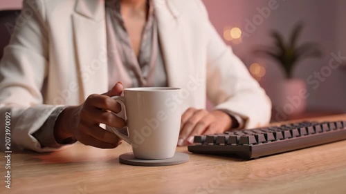A Professional Woman Enjoying Coffee While Working at Her Desk with a Computer Keyboard and Cozy Background Lighting in a Modern Office Setting