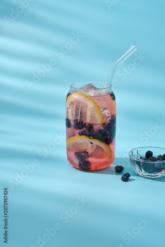 A refreshing drink, sparkling water with lemon, berries, and ice in a glass against a plain blue background.