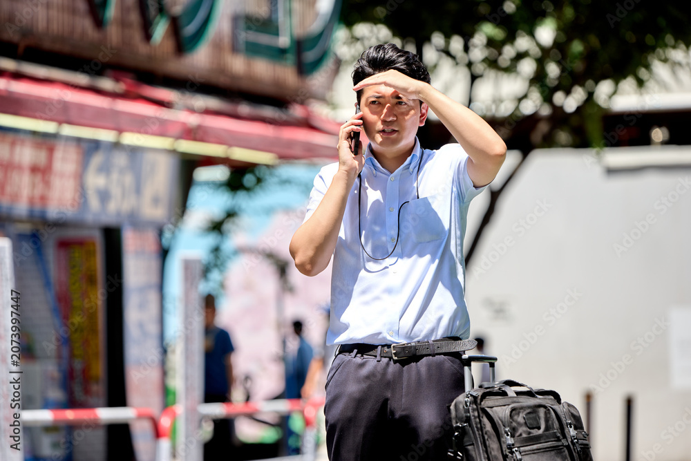 Fototapeta premium 屋外でスマートフォンで通話しながら日差しを遮る夏バテの仕事中の半袖の30代の日本人の男性の会社員