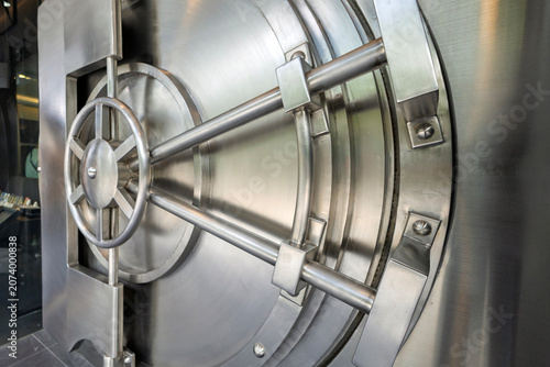 A close-up shot of a heavy-duty, circular stainless steel bank vault door showcasing its intricate mechanical locking wheel, security bolts, and solid metallic structure.