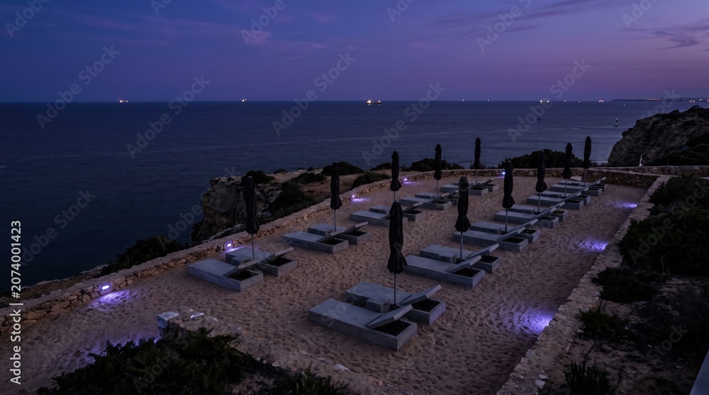 Obraz premium The scene shows a beach lounge area with many seats and umbrellas placed on the sand. Lights glow softly as the sun sets over the ocean, creating a relaxed atmosphere.