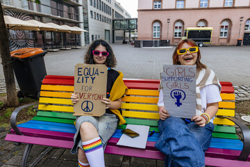 Friends outdoors sitting on rainbow bench holding equality signs