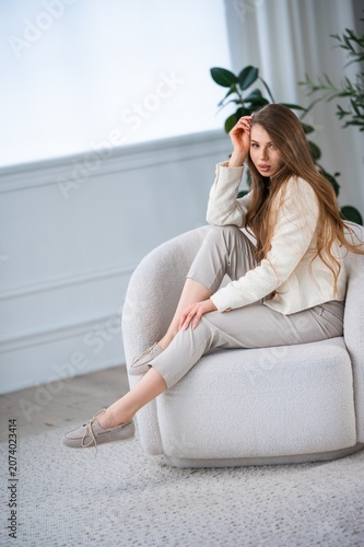 Young woman with long light brown hair seated on rounded textured armchair in bright minimalist interior, wearing neutral casual outfit and posing with calm relaxed expression