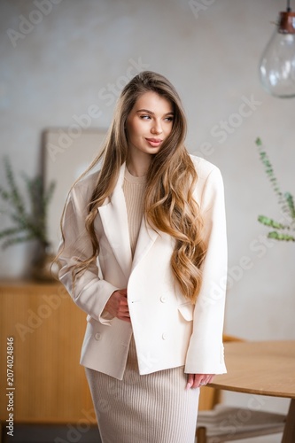 Young woman in cream blazer and ribbed knit dress posing indoors with long wavy hair, soft smile, neutral beige backdrop, modern pendant light and relaxed contemporary style