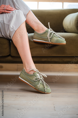 Woman in sage green sneakers sitting on olive sofa with crossed legs, casual pose, bright indoor setting, soft natural light, neutral dress hem and relaxed home styling