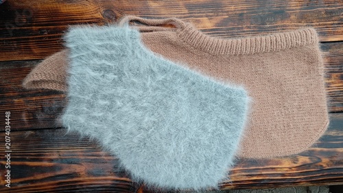 Horizontal flat lay of two cozy handmade winter hoods on a dark rustic wooden background. Fluffy light grey angora hood and beige wool balaclava. Warm winter accessories