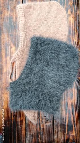 Vertical photo of handmade knitted balaclava hoods on a rustic wooden table. Fuzzy grey mohair and beige wool winter headwear