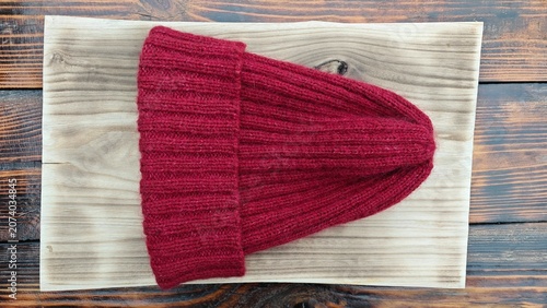 Horizontal flat lay of a red ribbed knitted beanie hat on a light wooden board. Handmade winter wool cap on a rustic tabletop