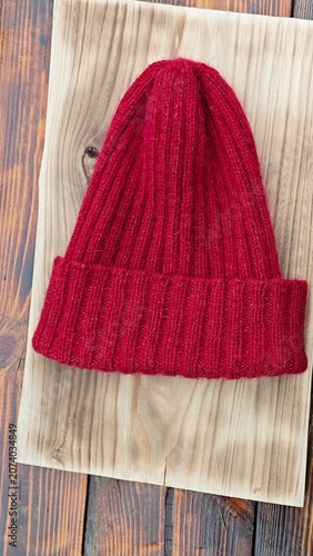 Vertical photo of a red ribbed knitted beanie hat on a light wooden board. Handmade winter wool cap on a rustic tabletop