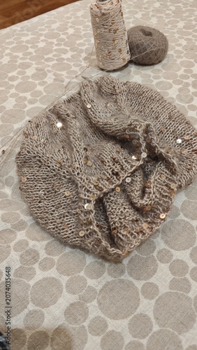Beige knitted scarf with gold sequins on patterned fabric. Handmade winter accessory with yarn ball and thread spool