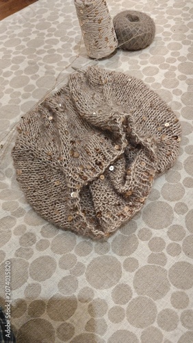 Close up of beige knitted scarf with gold sequins. Handmade winter accessory with yarn ball and thread spool