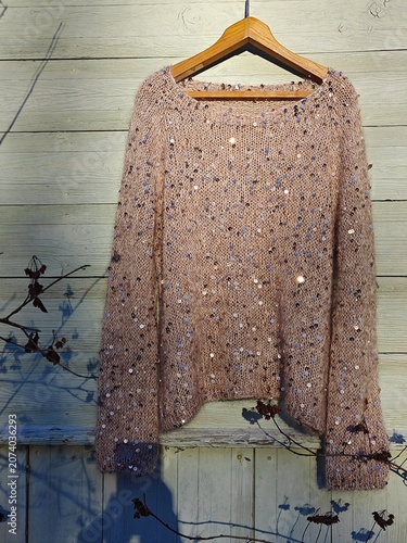 Handmade beige knitted sweater with sequins hanging on a wooden hanger. Finished garment displayed against a rustic light green wooden wall