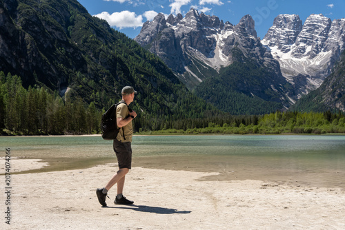 Male tourist with backpack walking on the sandy beach of Lake Landro and looking at majestic rocky mountain peaks, solo active hiking and summer wilderness exploration concept