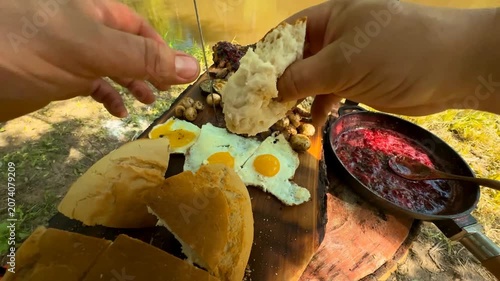 A hearty spread of rustic food prepared outdoors includes roasted meat fried eggs roasted potatoes and fresh bread enjoyed near a tranquil riverbank during a sunny summer day