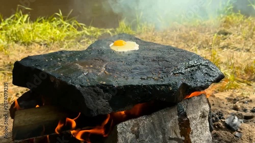 A person cracks and fries three eggs on a flat heated rock placed above a campfire near a river bank in a rustic natural setting under bright daylight hours
