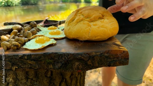 A rustic wooden board holds a hearty outdoor meal with grilled meat, fried eggs, roasted mushrooms, and sliced bread prepared near a calm river during a sunny summer day.