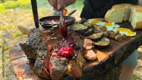 A rustic wooden board features a grilled tomahawk steak topped with tart cranberry sauce alongside fried eggs, roasted eggplant, and mushrooms in a serene riverside setting.
