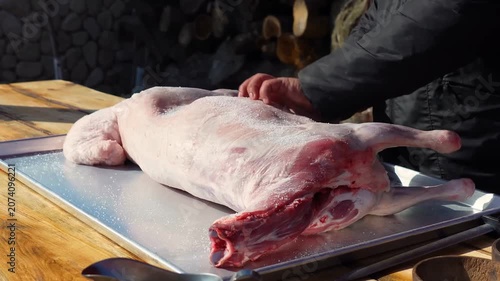 A whole raw lamb carcass is seasoned by hand with salt, pepper, and paprika on a large metal tray placed on a wooden outdoor table to prepare the meat for a traditional slow cooking process