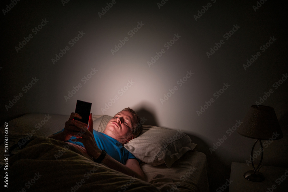 Fototapeta premium Man experiencing insomnia using smartphone in dark bed