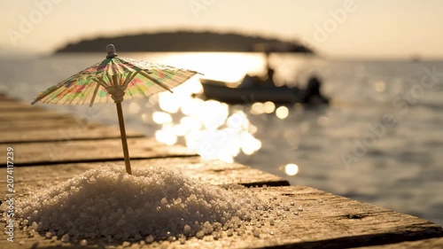 A vibrant beach scene featuring a colorful umbrella and salt scattered atop a wooden pier, with a picturesque sunset and boats gracefully drifting in the background.