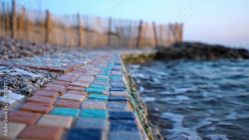 A serene coastal pathway adorned with colorful mosaic tiles, bordered by a gentle shoreline and a wooden fence, under the tranquil evening sky.