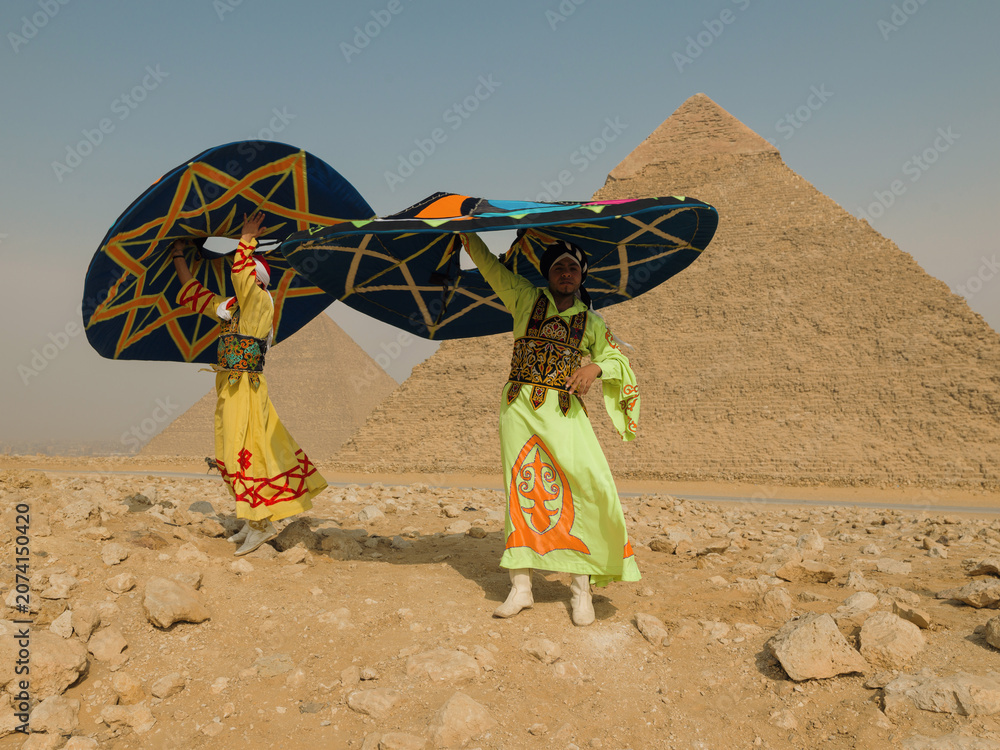 Fototapeta premium Two traditional folk dancers spin large colorful hats in vivid costumes before the Giza pyramids in Egypt celebrating cultural performance and tourism