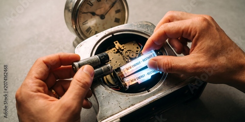 Person replacing old worn out batteries with glowing blue new energy cells inside vintage mechanical silver alarm clock on grey table.