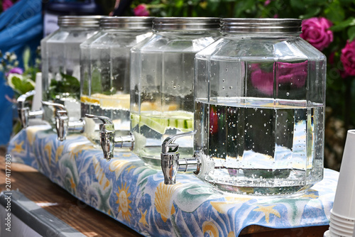 Large glass jar dispensers with tap faucets, filled with fresh water and some ingredients like lemon, cucumber or mint, to refresh visitors at a neighborhood street festival, copy space