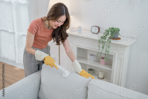 Woman cleaning sofa glove brush living room home dust cushion fabric, smiling woman cleaning fabric sofa cushion with brush and glove bright living room, tidy home interior with plant and white