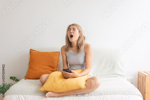 Tired young woman yawning widely on a comfortable sofa while holding her smartphone at home.