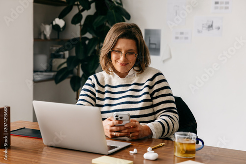 Obraz na plátne At her desk, the woman balances smartphone tasks with laptop work in a home offi