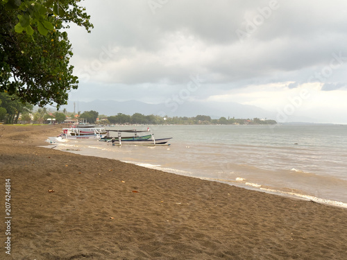 Traditional outrigger boats moored along the dark sandy shore of Lovina Beach under a cloudy sky, Buleleng, North Bali, Indonesia.