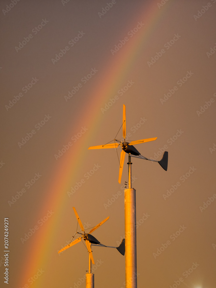 Naklejka premium features two wind turbines silhouetted against a golden hour sky with a rainbow.