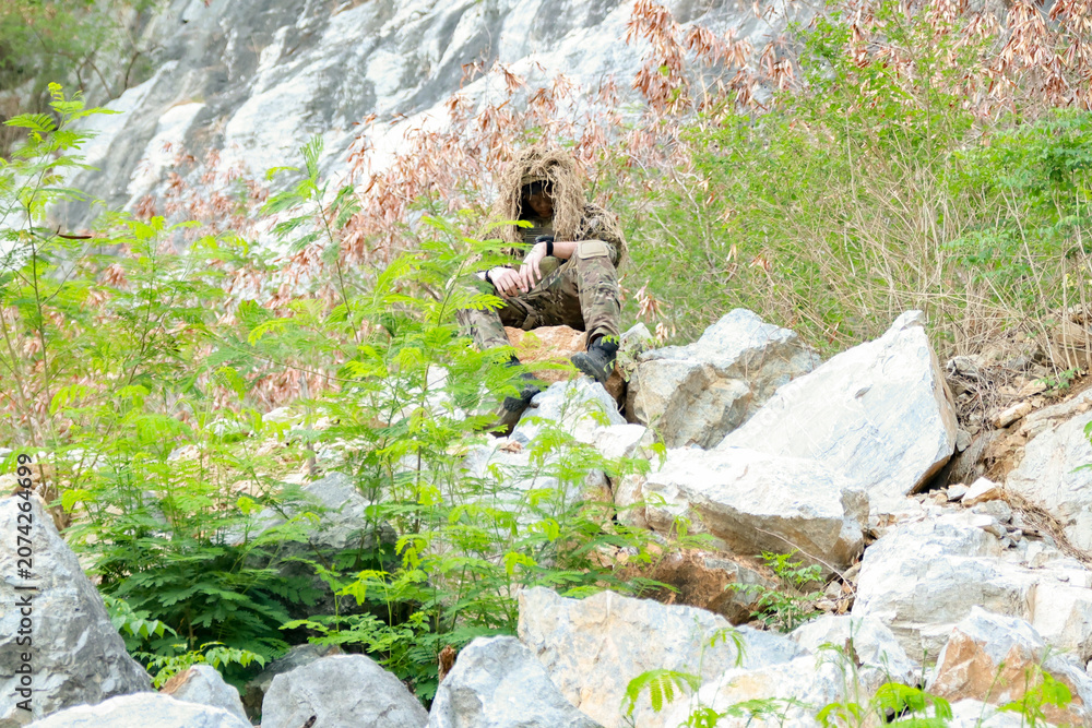 Fototapeta premium Male soldier in camouflage uniform sitting on a rock on the edge high cliff in a mountainous area closely observing the border situation.