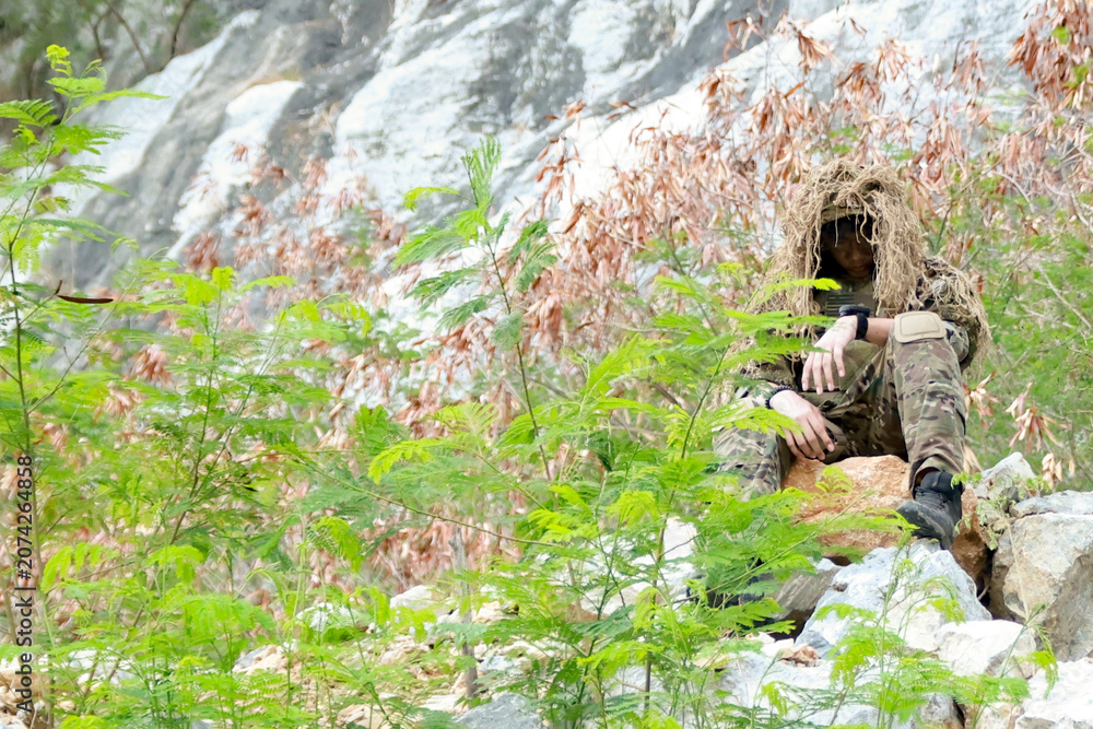 Fototapeta premium Male soldier in camouflage uniform sitting on a rock on the edge high cliff in a mountainous area closely observing the border situation.