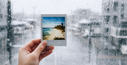 Mano con fotografia istantanea di paesaggio tropicale, sfondo con superficie vetrata coperta di pioggia con vista su panorama invernale