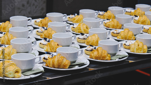 Rows of coffee break sets with croissants and tarts on plates for seminar conference catering.
