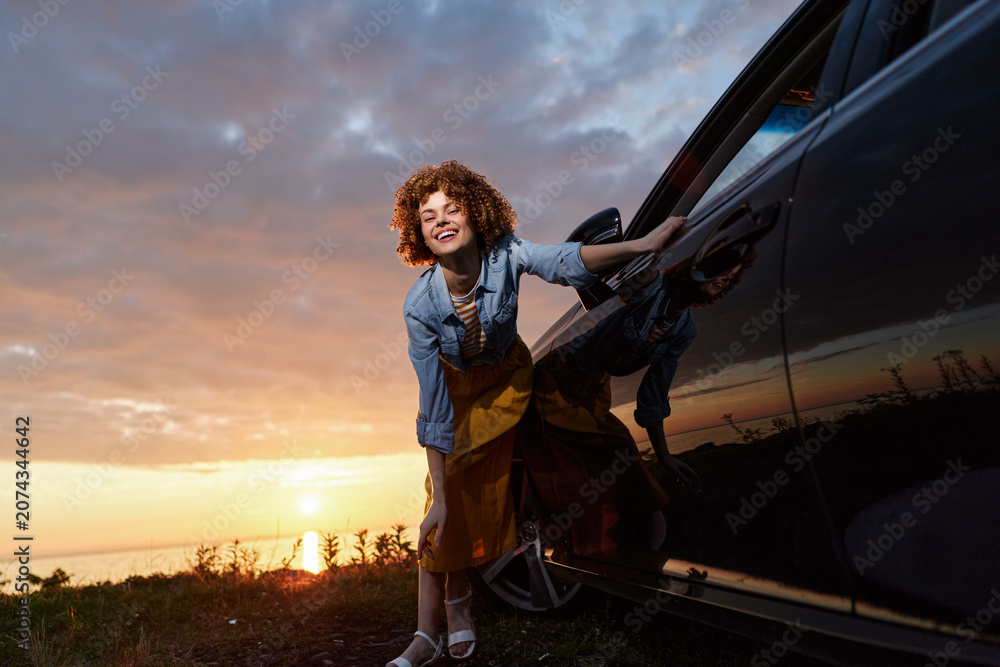 Fototapeta premium Smiling woman with curly hair standing beside a car at sunset. Casual denim shirt and dress convey outdoor lifestyle, warmth, energy, and everyday freedom. Natural light creates a bright, candid,