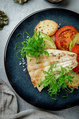 Close-up seafood dish with grilled white fish fillet, roasted vegetables, fresh herbs, sea salt and black plate on grey table