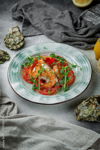 Vertical seafood appetizer with grilled shrimp, tomato slices, creamy cheese, microgreens, lemon and oyster shells on textured grey table