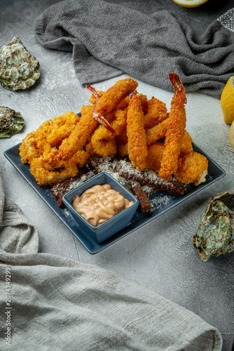 Vertical crispy fried seafood platter with breaded shrimp, calamari rings, dipping sauce and lemon on a grey textured restaurant table