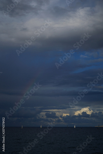 Vertical shot of a fleet of small sailing dinghies on the horizon under a dramatic, stormy sky in Crete, with a faint rainbow appearing over the dark Mediterranean sea.